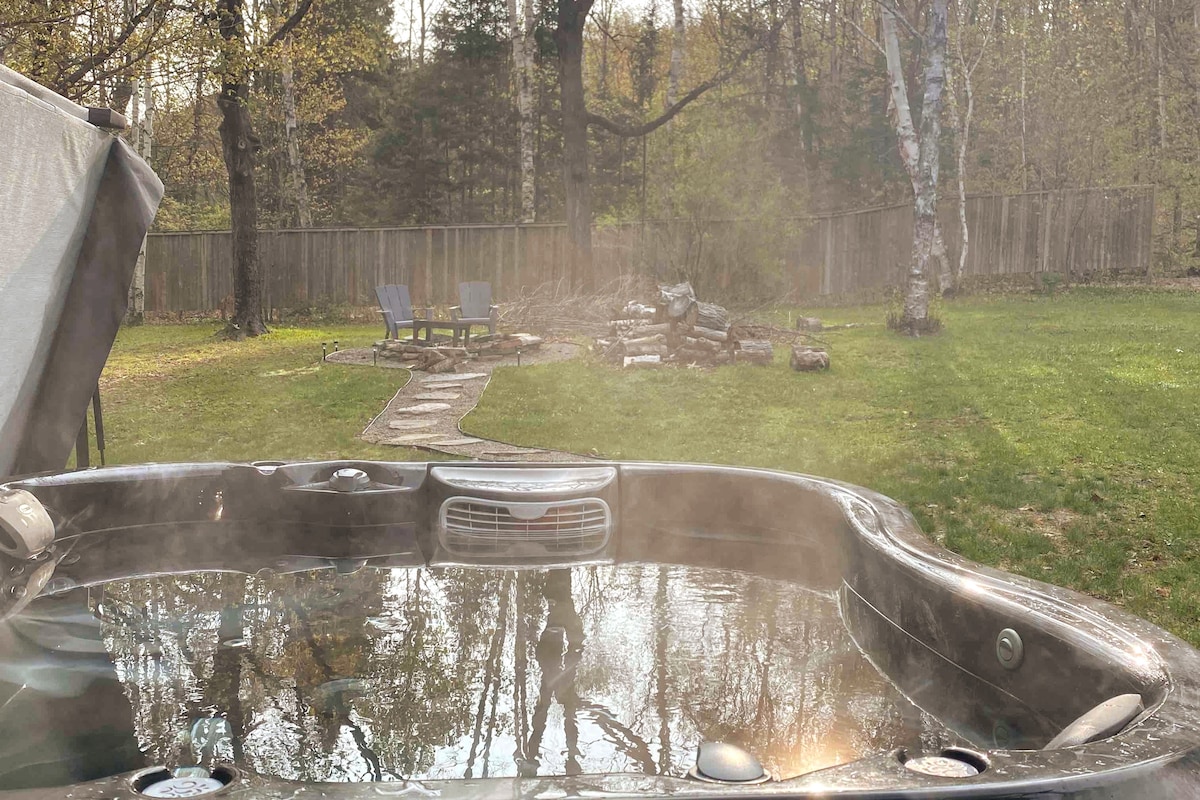 A steaming hot tub sits in the foreground, reflecting trees and the sky. In the background, a pathway lined with stones leads to a fire pit surrounded by logs, framed by lush green grass and tall trees.