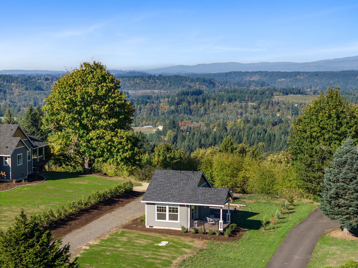 A craftsman-style guesthouse with a dark roof is situated on a spacious green parcel, surrounded by trees and distant mountain views. The outdoor area features a gravel driveway leading up to the home, highlighting the peaceful countryside setting.