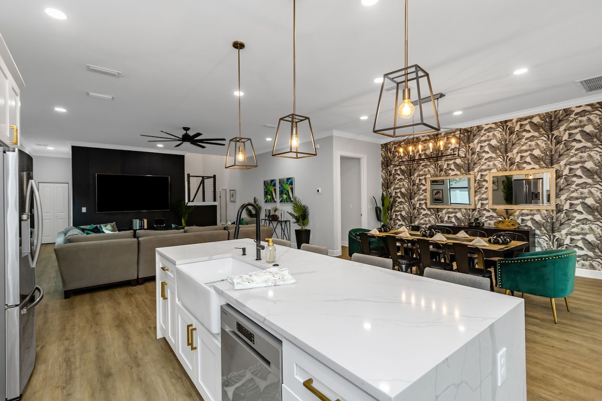 An open-concept kitchen and dining area showcases modern pendant lighting above a large island with a double sink. The kitchen is equipped with stainless steel appliances and white cabinetry. The living space, visible in the background, includes comfortable seating and a large wall-mounted television.