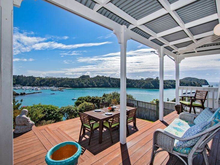 An outdoor deck with natural wood flooring offers a spacious dining table surrounded by chairs. A scenic view of the turquoise harbor is framed by white support beams, with soft clouds lazily drifting through a bright blue sky.