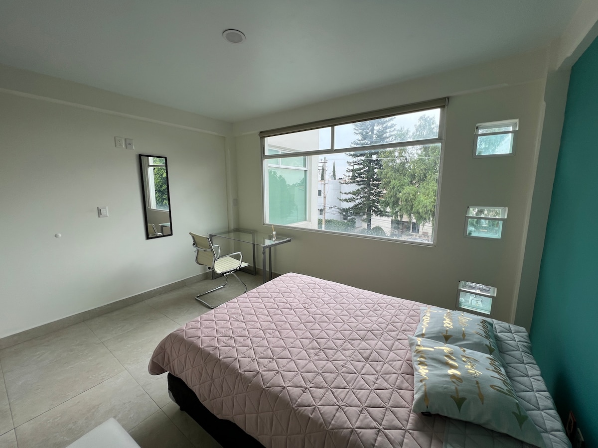 A bright bedroom is highlighted by large windows allowing natural light to fill the space. A spacious bed is dressed in soft linens, accompanied by decorative pillows. A sleek desk and chair are positioned nearby, and a mirror is mounted on the wall, reflecting the room's tranquil design.