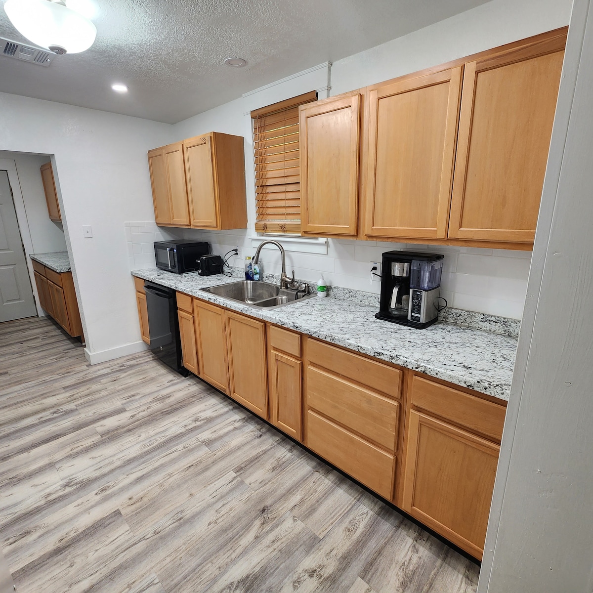 The kitchen features wooden cabinetry with a granite countertop and stainless steel appliances. A double sink is located between a coffee maker and a microwave. Natural light enters through a nearby window, illuminating the light-colored flooring.