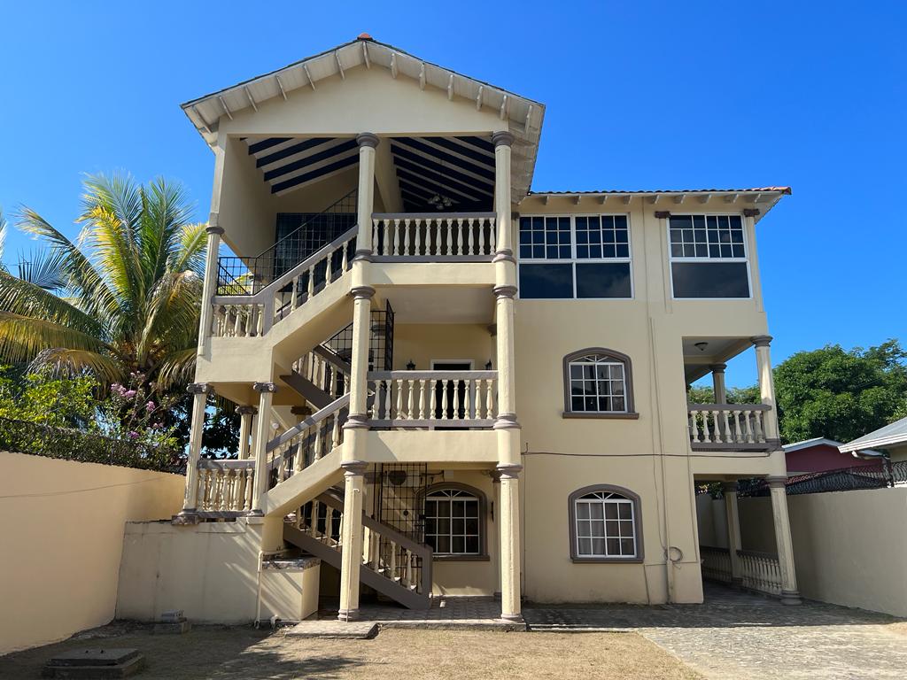 A three-story building is featured, showcasing a structured design with decorative pillars and a balcony. Surrounding greenery includes palm trees, while multiple windows allow natural light to fill the space. A well-maintained outdoor area is visible, providing a sense of openness.