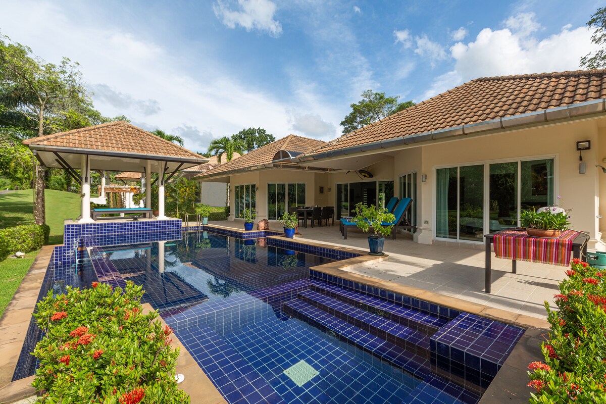 The villa features a private saltwater infinity pool, with a jacuzzi at one end and sun loungers positioned nearby. A covered outdoor dining area provides shade, and lush greenery surrounds the patio, creating a serene atmosphere with views of the golf course.