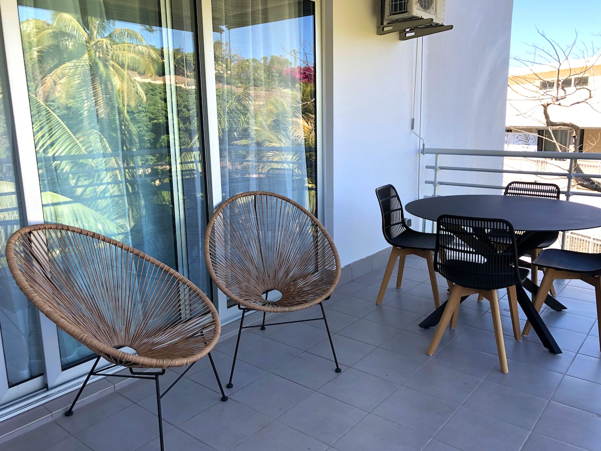 A balcony area is featured with two woven chairs and a round dining table surrounded by four chairs. Large glass doors provide access to the space, while greenery is visible in the background, creating a serene outdoor ambiance.