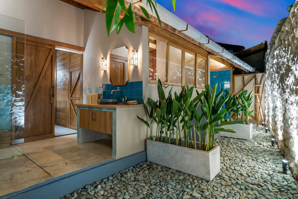An outdoor view of the villa's entrance showcases a modern design with natural elements. Lush greenery is framed by smooth pebbles, while soft lighting highlights the wooden doors and blue tiled accents. The space blends indoor comfort with outdoor tranquility.