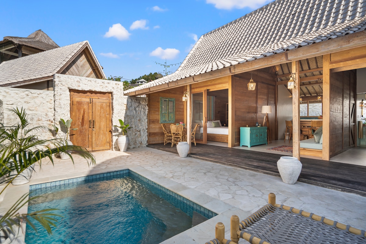 A tranquil outdoor space features a private plunge pool surrounded by natural stone tiles. The villa's open structure showcases elegant wooden beams, with inviting spaces visible through expansive doors. A comfortable seating area and lush greenery add to the serene atmosphere.