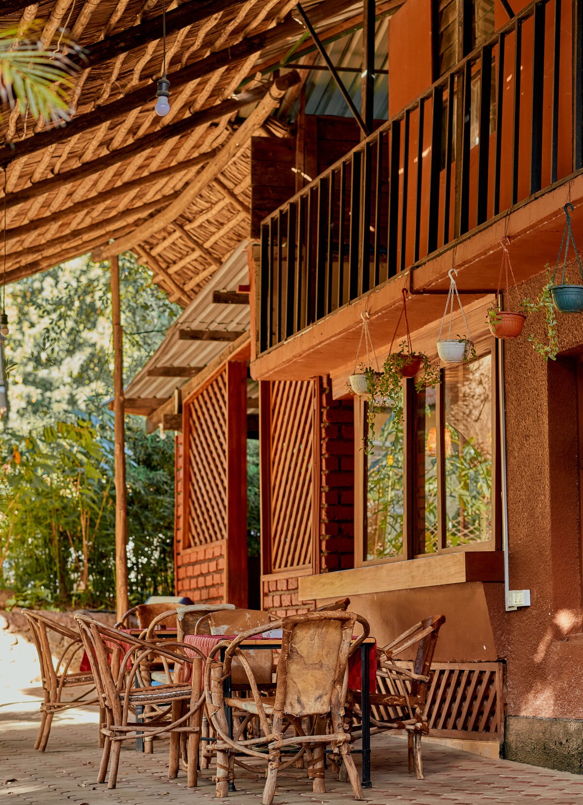 An outdoor seating area is displayed with a wooden table surrounded by several woven chairs. The shaded area is enhanced by a thatched roof, with hanging potted plants adding a touch of greenery. The structure features wooden beams and large windows.