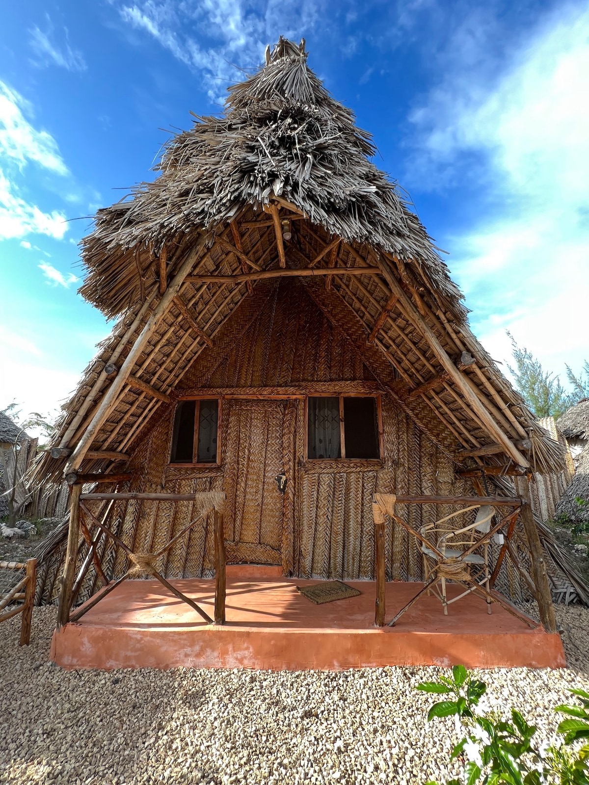 A charming bungalow features a thatched roof and wooden accents, surrounded by natural elements. The front porch has a small seating area with a chair, and the entrance is framed by two windows, enhancing the rustic appeal of the structure.