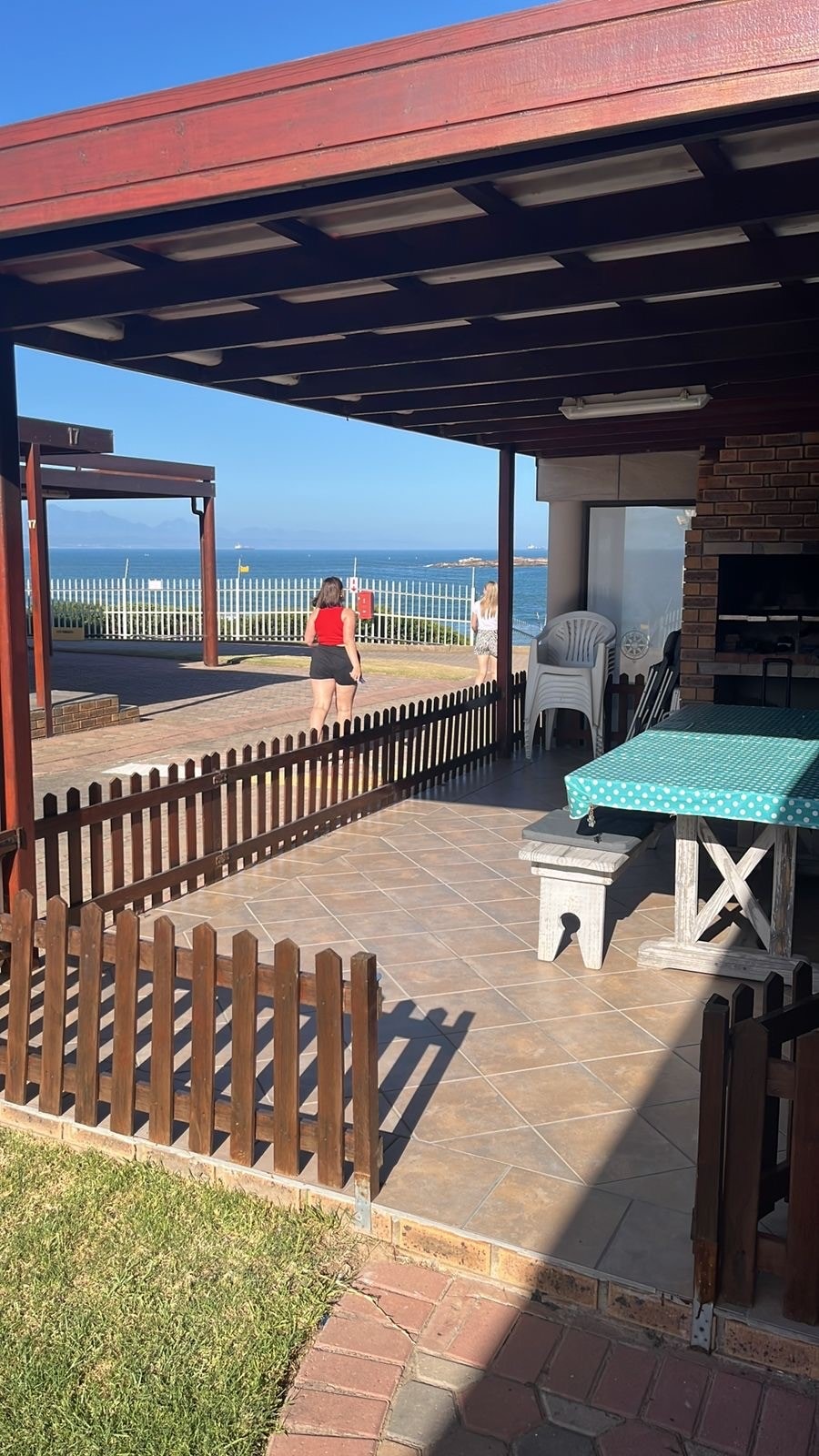 An outdoor area features a covered patio with a blue tablecloth atop a sturdy table. A fenced section of the yard is visible, alongside a grassy area. The ocean can be seen in the background, with a couple of individuals enjoying the nearby space.
