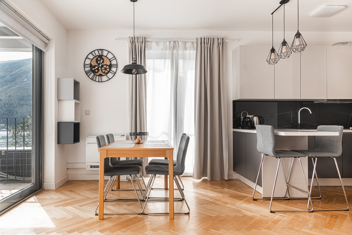 A dining area features a wooden table accompanied by four gray chairs. The kitchen is visible with sleek black cabinets and modern appliances. Natural light enters through large glass doors, illuminating the space, while a wall clock adds a decorative touch.