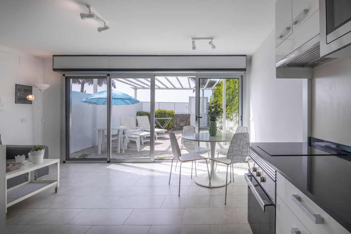 A modern living area is visible, featuring a dining table surrounded by four white chairs. Natural light enters through large sliding glass doors, leading to a private outdoor terrace equipped with seating and an umbrella. A minimalist kitchen is seen in the corner.