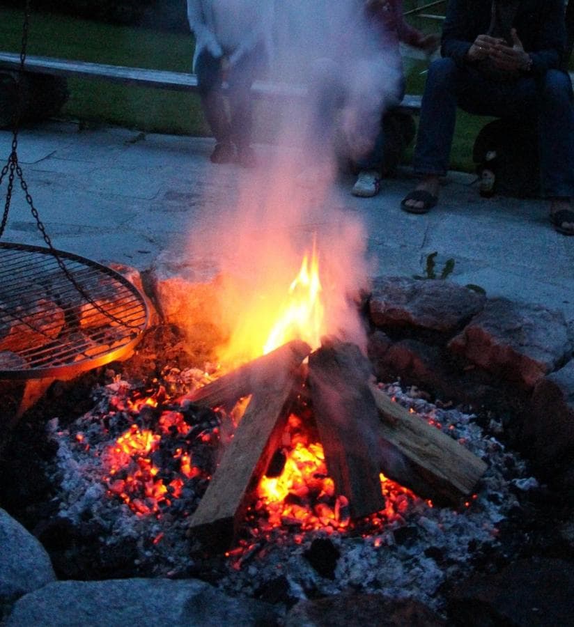 A circular stone fire pit is filled with burning logs, emitting flames and smoke into the evening air. Nearby, people are seated on a bench, partially visible, creating an inviting communal space for gathering around the warmth of the fire.
