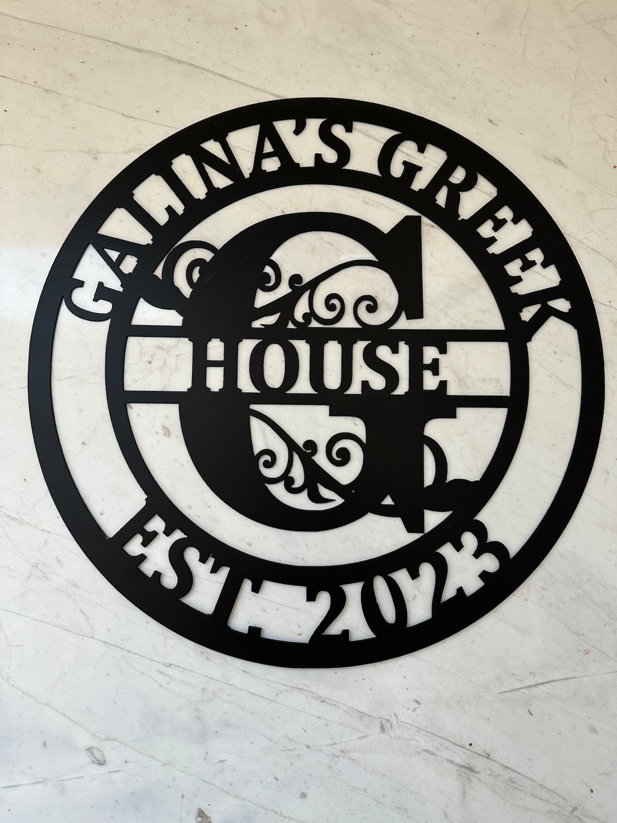 A decorative metal sign featuring the name 'Galina's Greek House' prominently displayed. The design incorporates intricate scrollwork around the letter 'G' and includes the establishment year, 'EST 2023.' The sign is set against a light, textured marble background.