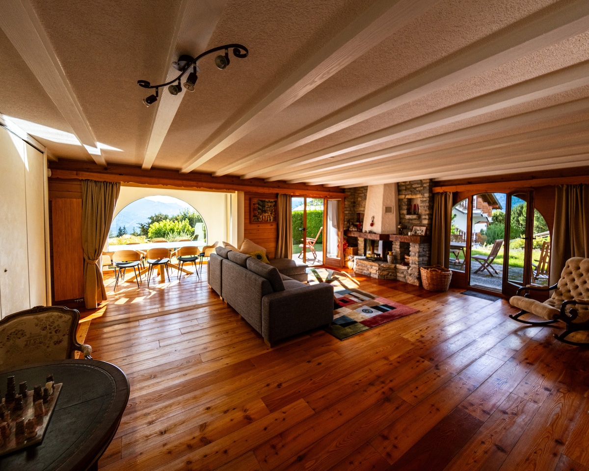 The spacious living area features wooden beams and flooring, illuminated by natural light. A cozy sofa is situated near a stone fireplace, creating a warm ambiance. Large windows offer views of the garden and mountains, with a dining area visible to one side.