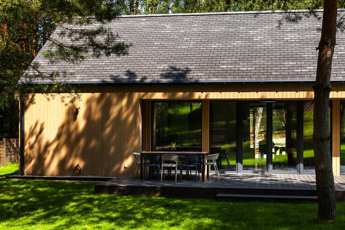 The exterior of a modern house is depicted, featuring wooden cladding and large glass doors that open to a deck. A spacious outdoor dining area is visible, along with a lush green lawn surrounded by trees.