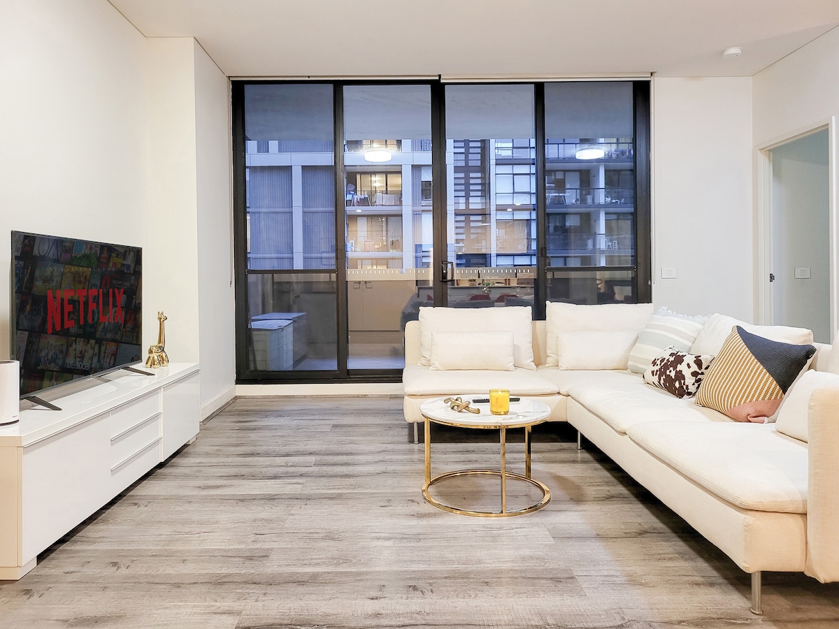 A modern living area features a spacious, light-colored sectional sofa positioned adjacent to a sleek media console with a TV. Large glass doors provide views of the external area, while natural light enhances the wood-like flooring. A circular coffee table sits in the center of the space.