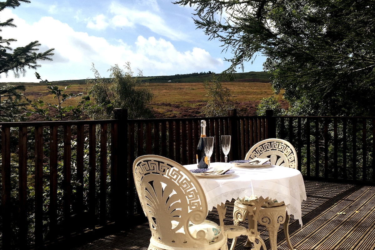 A small round table is set for two on a wooden decking area, accompanied by elegant white chairs. A bottle of champagne and two glasses are arranged on the table. The surrounding trees provide a natural frame, while a scenic view of the moorland stretches in the background.