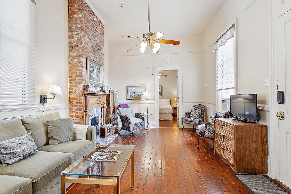 The living area is equipped with a comfortable sectional sofa and features natural wood flooring. A wall of exposed brick adds character, while a ceiling fan overhead provides ventilation. A glass coffee table sits in front, and a small television is positioned on a wooden dresser.
