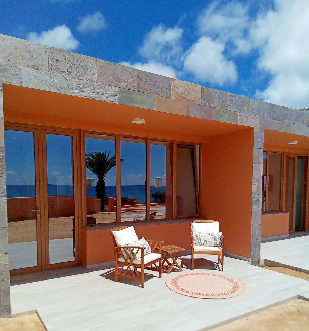 An exterior view showcases a modern building with stone and warm-toned walls. Two wooden chairs with light cushions sit on a porch, accompanied by a round rug. Large windows reflect the bright blue sky and a glimpse of the ocean can be seen in the background.