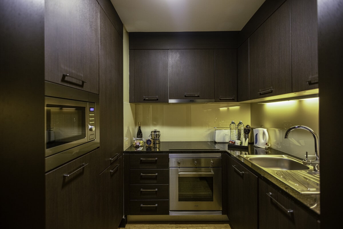 A well-equipped kitchenette features dark wooden cabinetry and a granite countertop. Appliances include a microwave, kettle, and oven, while organized utensils and glassware are neatly arranged. Under-cabinet lighting illuminates the space, enhancing its functionality and modern design.