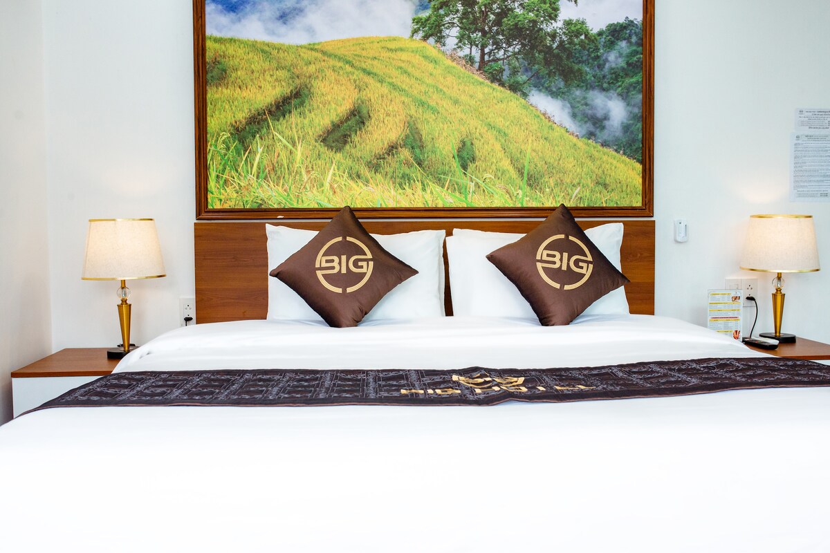 A spacious bed is dressed in crisp white linens, accented by decorative pillows featuring the BIG logo. Soft bedside lamps provide warm lighting, while a large artwork depicting lush green rice terraces hangs above the headboard, enhancing the serene atmosphere of the room.
