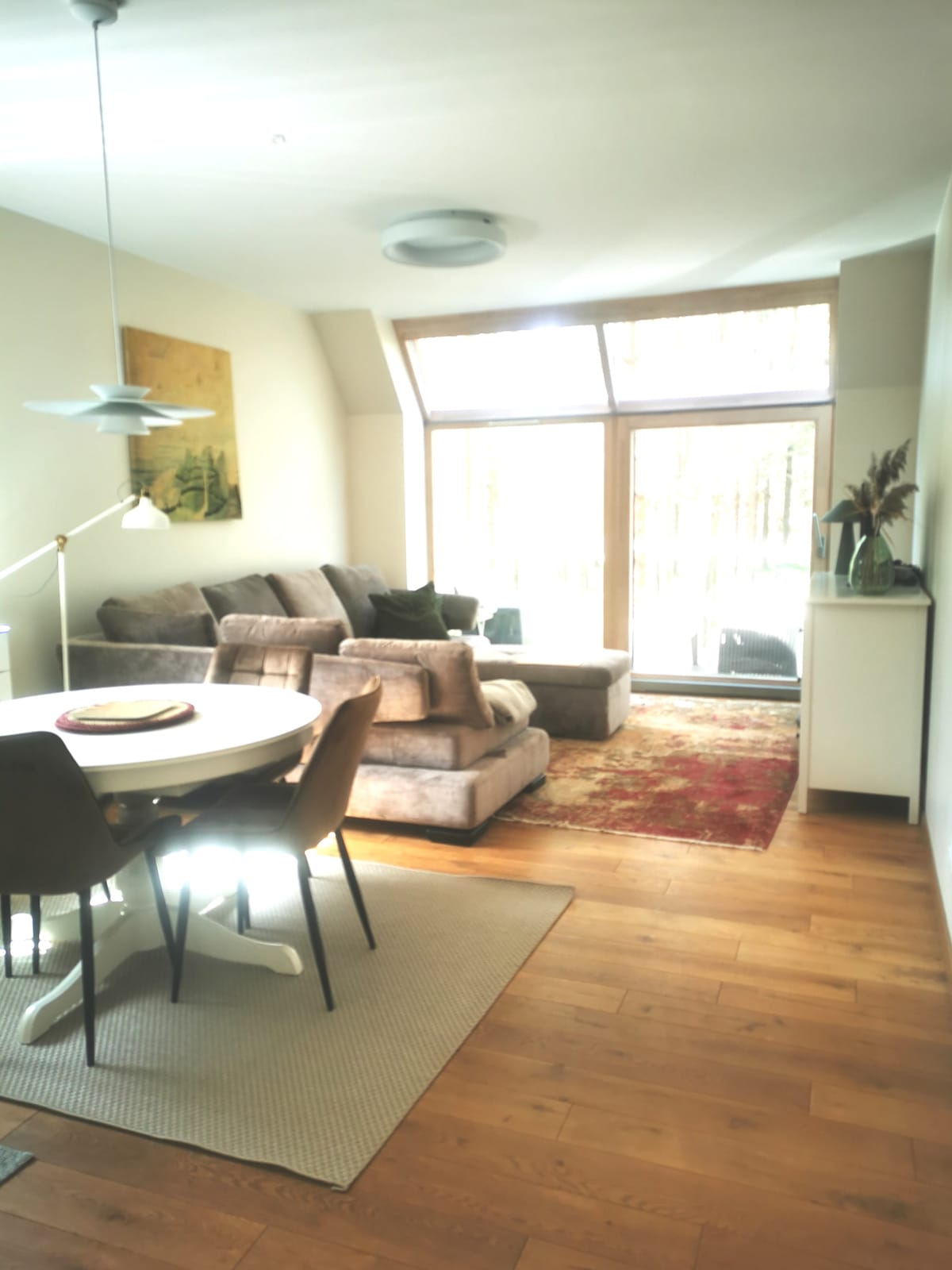 A spacious living area features a comfortable sectional sofa and a round dining table with chairs. Natural light filters through large glass doors, leading to a balcony. Warm wood flooring adds to the inviting atmosphere, complemented by a soft area rug.