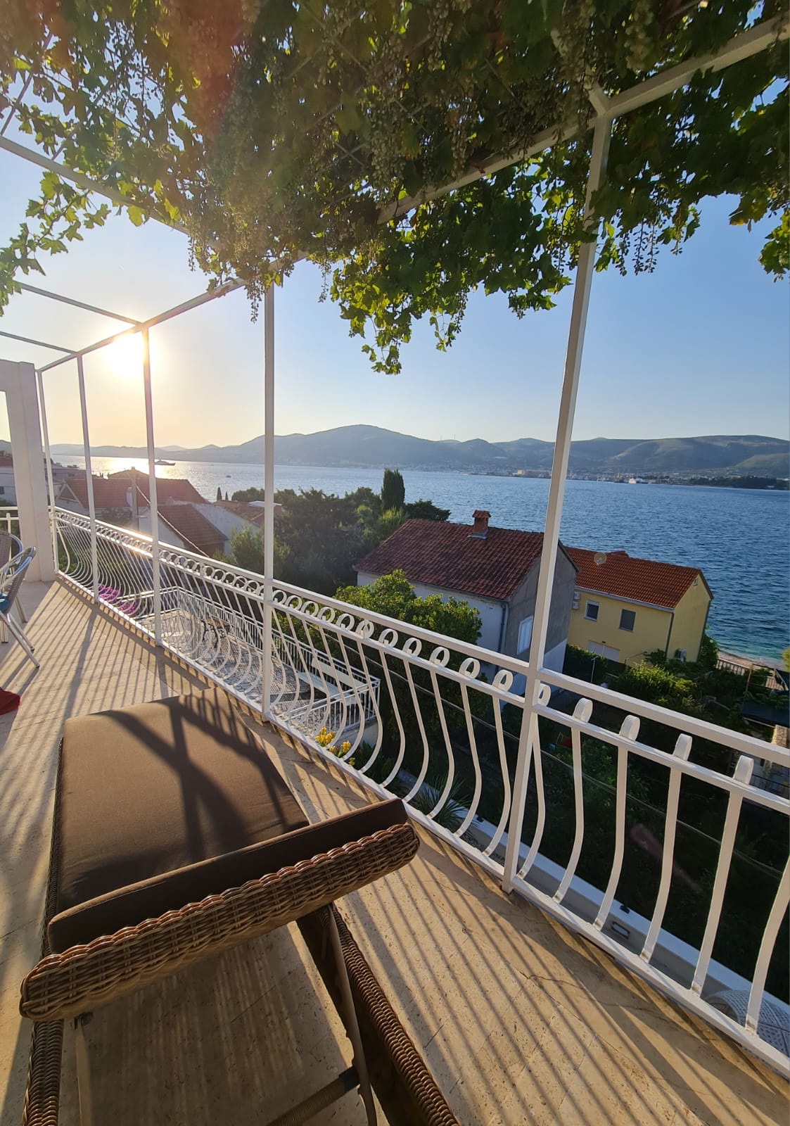 A spacious balcony is presented, featuring a comfortable seating area overlooking the sparkling water. The sun sets over the distant hills, creating a warm glow. Lush greenery partially frames the scene, enhancing the peaceful coastal view of the surrounding area.