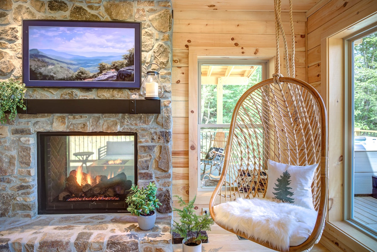 A stone fireplace is set against a wooden wall, providing warmth with a gentle flame. A modern hanging chair is positioned nearby, featuring a cozy cushion adorned with a tree design. Large windows allow natural light to fill the space while showcasing views of the surrounding outdoors.