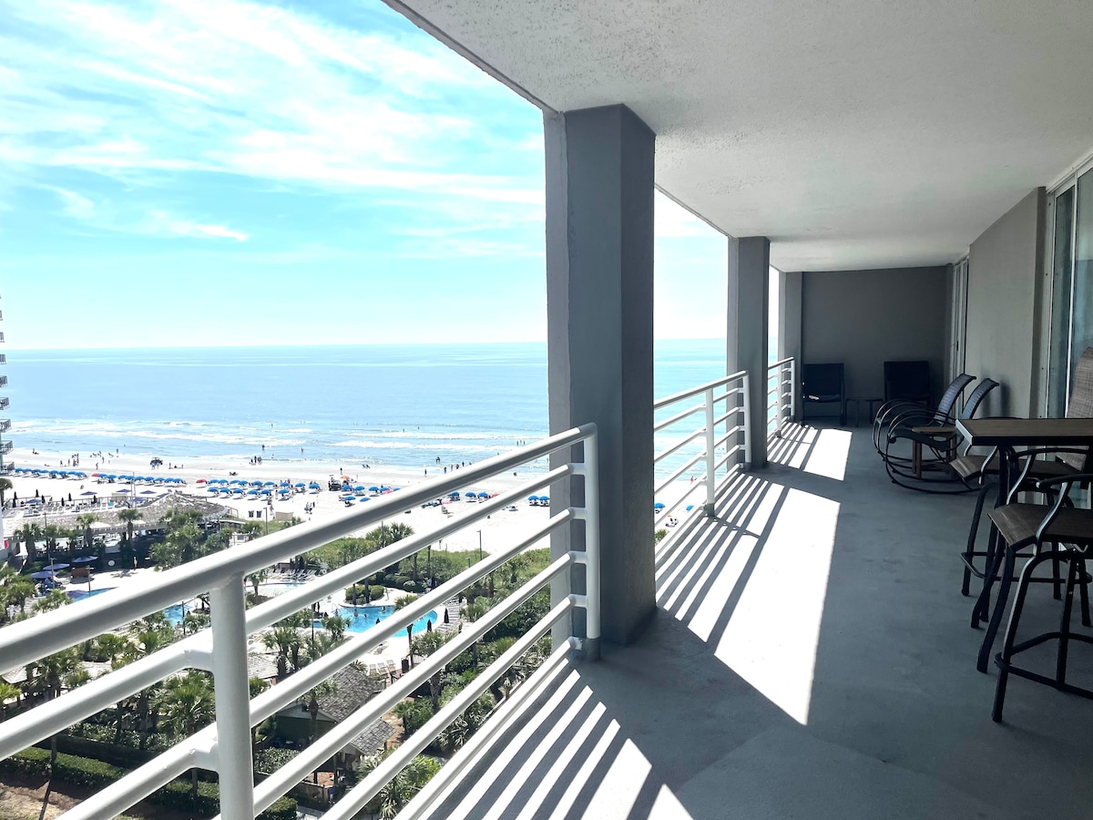 A spacious balcony offers unobstructed ocean views. Comfortable seating arrangements can be seen, alongside a dining table. The gentle sunlight casts distinct shadows across the surface, highlighting the coastal landscape below, characterized by sandy beaches and inviting blue waters.