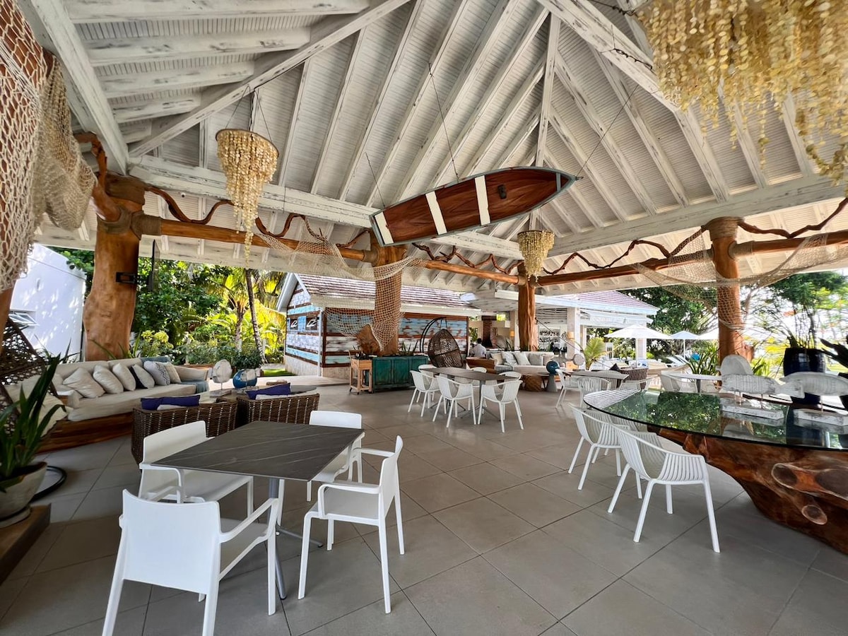 An open-air social area features a high vaulted ceiling with wooden beams. Natural light floods the space, showcasing a mix of seating options including a dining table and lounge areas with plush cushions. Unique light fixtures hang down, enhancing the inviting ambiance.