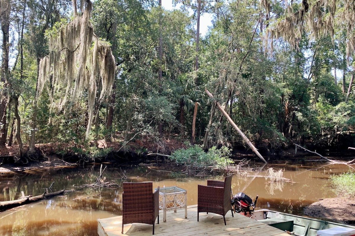 A private floating dock is surrounded by lush greenery, with two wicker chairs positioned near a small table. Spanish moss hangs from nearby trees, providing shade over the tranquil waterway, where reflections of the surrounding nature are visible.