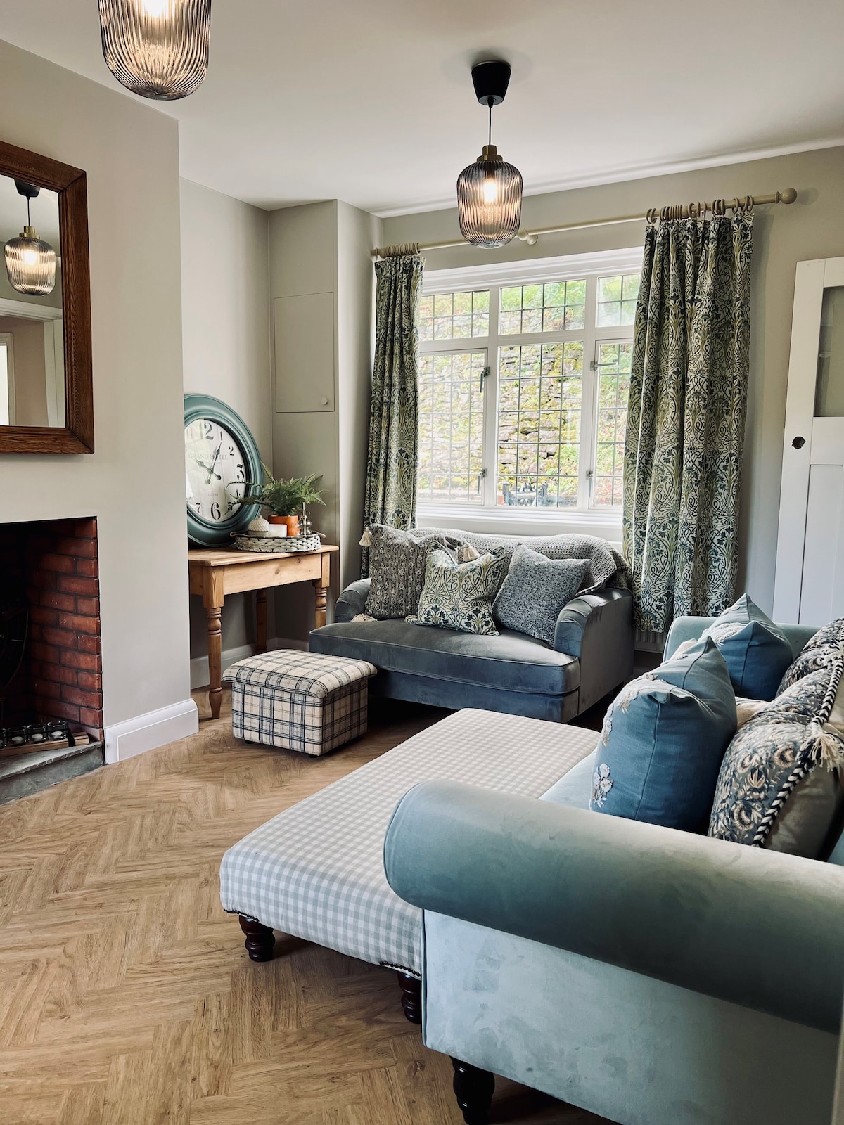 The inviting lounge area features a combination of soft seating, including a light blue sofa and a patterned armchair. Natural light enters through large windows dressed with patterned curtains. A charming clock and a small table are positioned near a classic fireplace, enhancing the relaxed atmosphere.