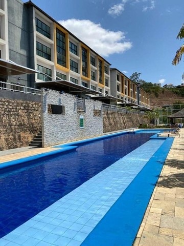 An outdoor swimming pool is set against a modern multi-story building. Clear blue water reflects the sky, while stone walls provide a textured backdrop. Sun loungers are positioned along the pool's edge, complemented by shaded areas created by nearby trees.