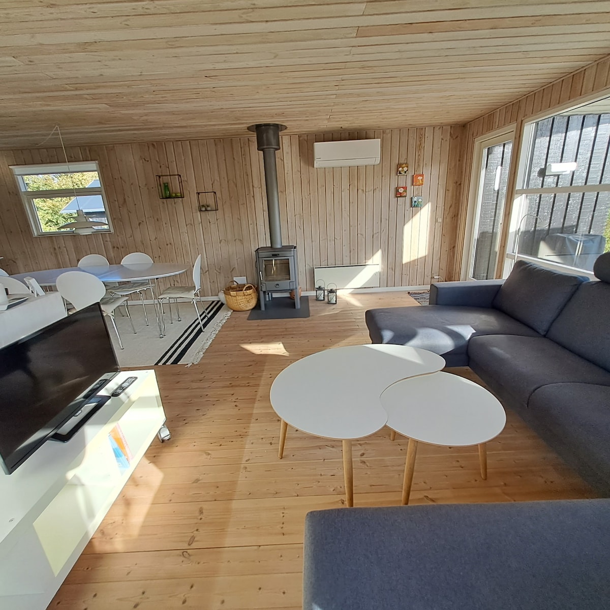 An open living space features wooden walls and a light wooden floor. A comfortable sofa faces a small coffee table, with a television nearby. A modern wood stove stands near large windows, allowing natural light to brighten the area. A dining table is visible in the background.