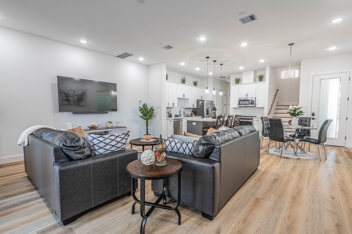 An open-concept living area features a modern kitchen and dining space, illuminated by recessed lighting. Comfortable leather sofas are arranged around a circular coffee table. A dining table for four is set nearby, creating a welcoming space for relaxation and dining.