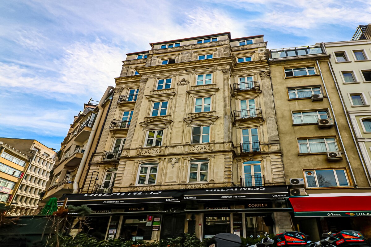 A historic building is observed from street level, showcasing intricate architectural details and several balconies. Large windows reflect the sky, while the ground floor features storefronts with signage. Surrounding greenery and parked motorcycles contribute to the lively urban atmosphere.