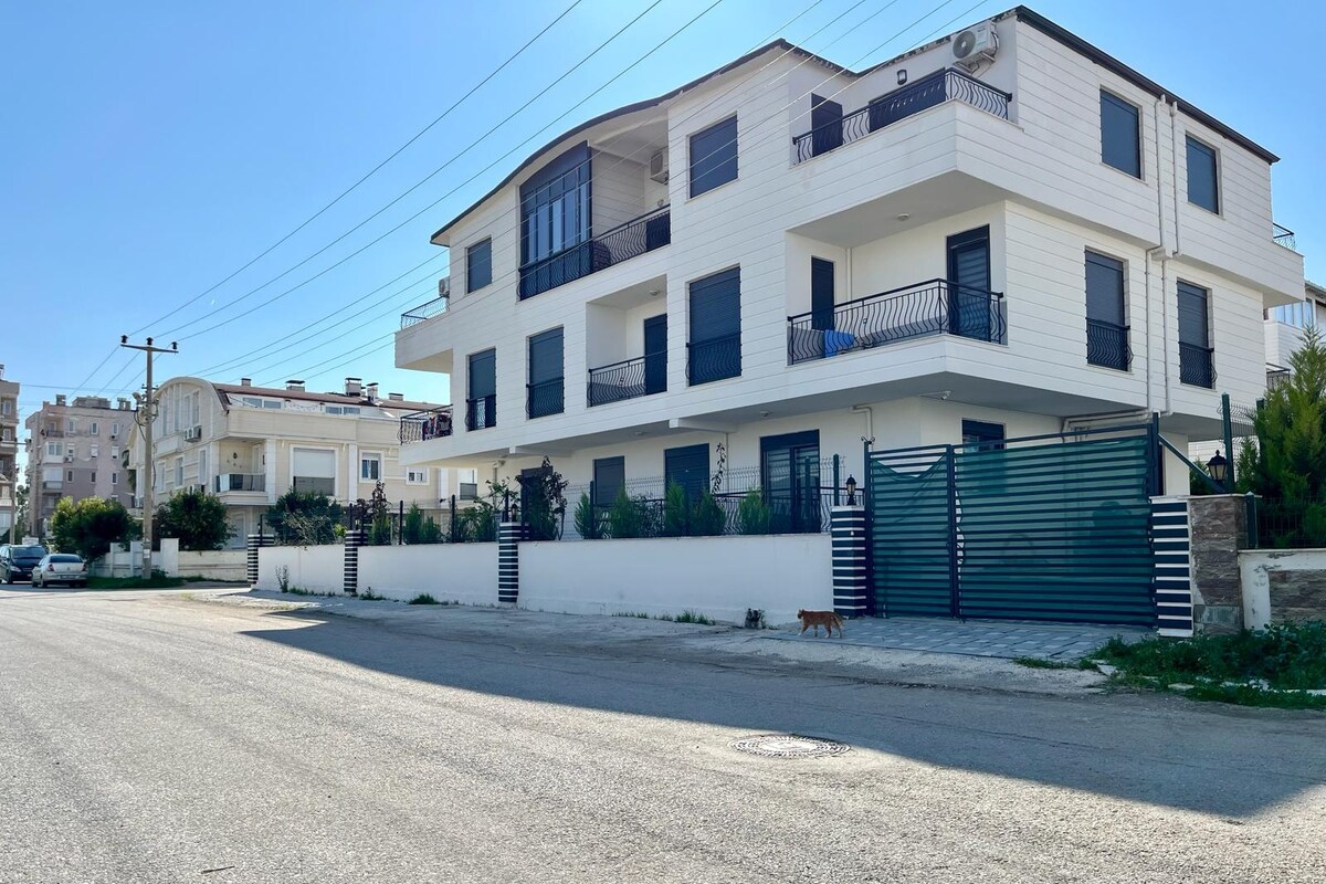 The exterior of a modern multi-unit building is presented, featuring white paneling and a mix of balconies. A well-maintained garden surrounds the entrance, which is secured by a green gate. The street scene indicates a quiet residential area with minimal traffic.