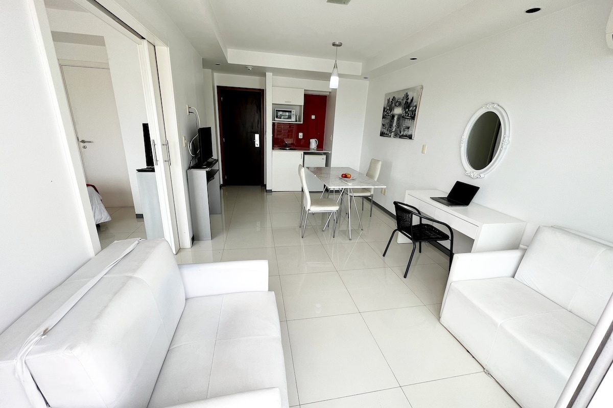 A modern living space features sleek, white furniture, including two armchairs and a sofa. A dining table with four chairs is set against a backdrop of bright red cabinetry. A small workspace with a laptop and contemporary decor completes the inviting atmosphere.