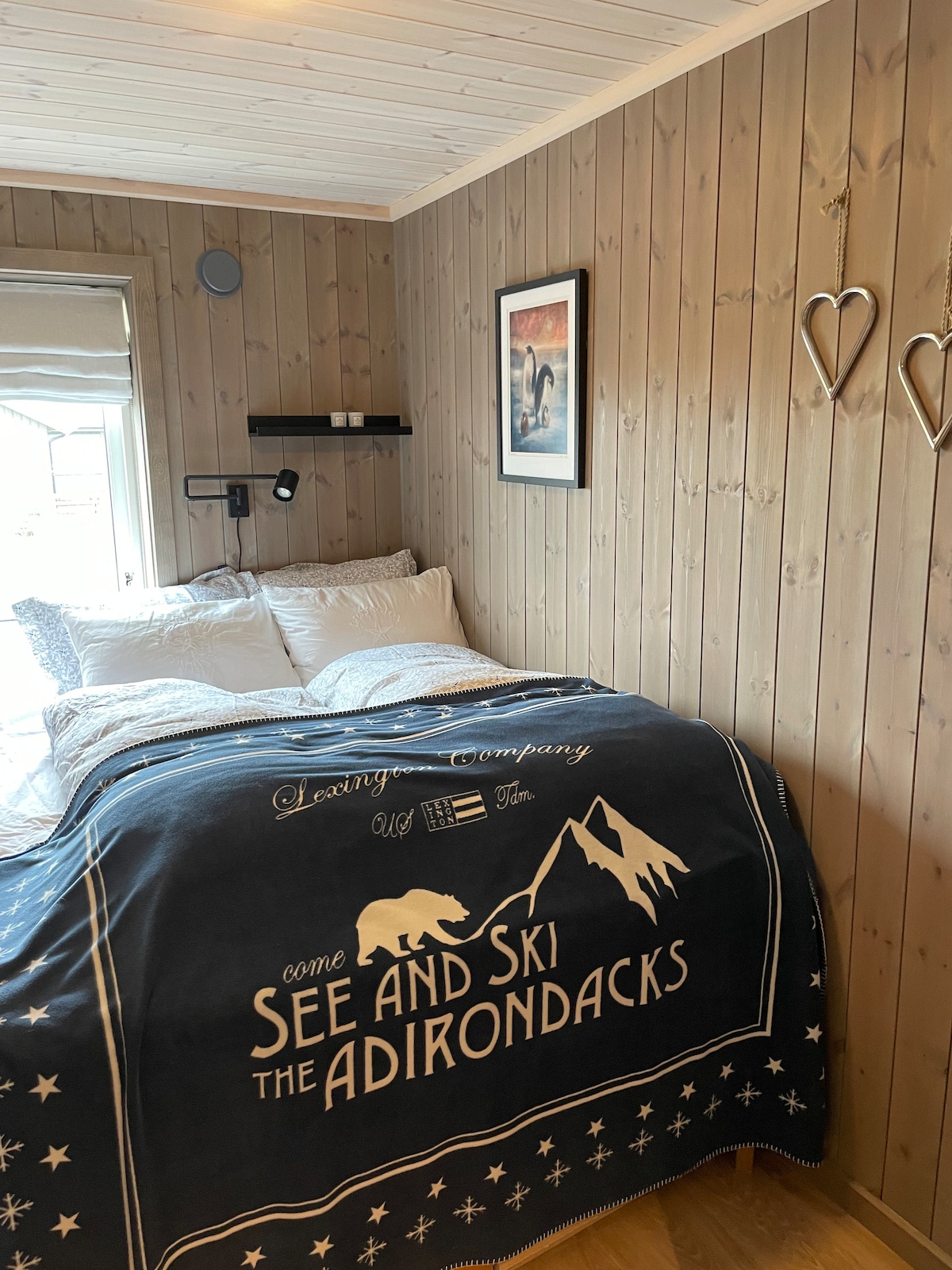 A cozy sleeping area features wooden walls and a large window that allows natural light to fill the space. A spacious bed is adorned with a patterned blanket. Simple decor, including a framed picture and wall-mounted shelves, contributes to a comfortable atmosphere.