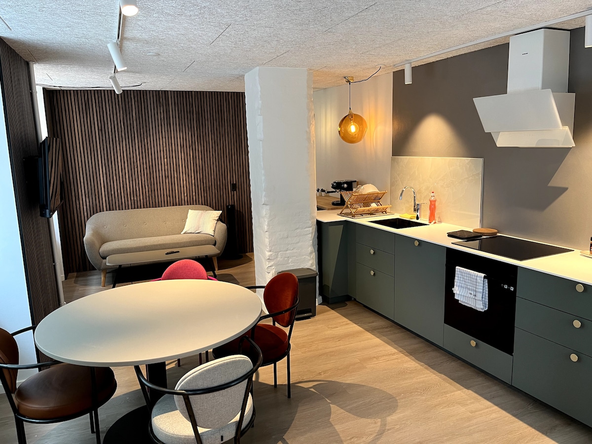 An open kitchen and dining area are displayed, featuring a round table surrounded by six chairs of various colors. A cozy sofa is positioned nearby, and a television is mounted on the wall. Modern cabinetry and appliances are visible in the kitchen space.