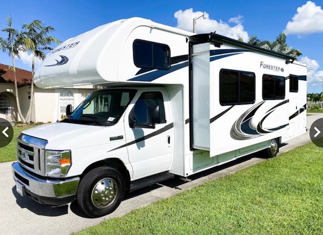 A spacious Class C motorhome sits on a well-maintained lawn, showcasing a sleek white exterior with stylish graphics. The vehicle features large windows for natural light, a prominent cab area, and an overhead sleeping compartment. Lush greenery surrounds the RV, enhancing its inviting appearance.