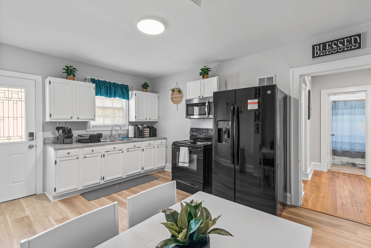 A fully equipped kitchen is presented, featuring black appliances, including a refrigerator and stove. White cabinets and countertops create a bright atmosphere. A dining table with a plant centerpiece is situated in the foreground, while natural light enters through the window with blue curtains.