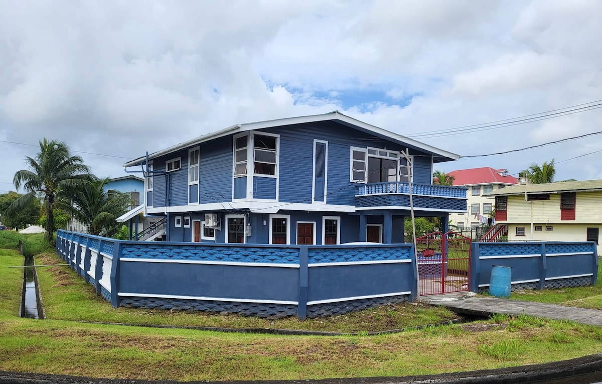 The exterior of the two-story house is showcased, featuring blue siding and multiple windows. A well-maintained yard surrounds the property, bordered by a decorative fence. Tropical palm trees and nearby buildings enhance the vibrant setting.