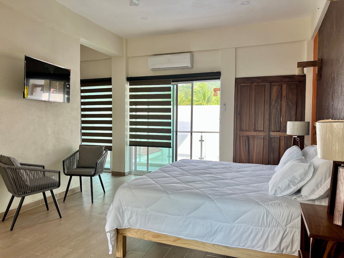 A spacious bedroom is presented with a large bed covered in white linens. Two accent chairs are placed near a window with modern blinds. A television is mounted on the wall, and a wooden cabinet adds warmth to the space.