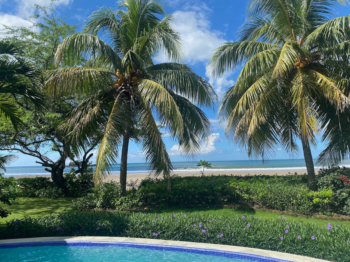 Lush palm trees frame the view of Marsella Beach, providing a serene backdrop to the private pool. The calm ocean can be seen in the distance, with a clear blue sky accentuating the tranquil atmosphere of the garden area.