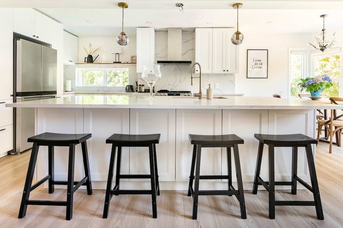 A modern kitchen features a large island with a sleek countertop, complemented by four black wooden stools. Natural light streams in through large windows, while minimalist cabinetry and stylish light fixtures add to the contemporary aesthetic.