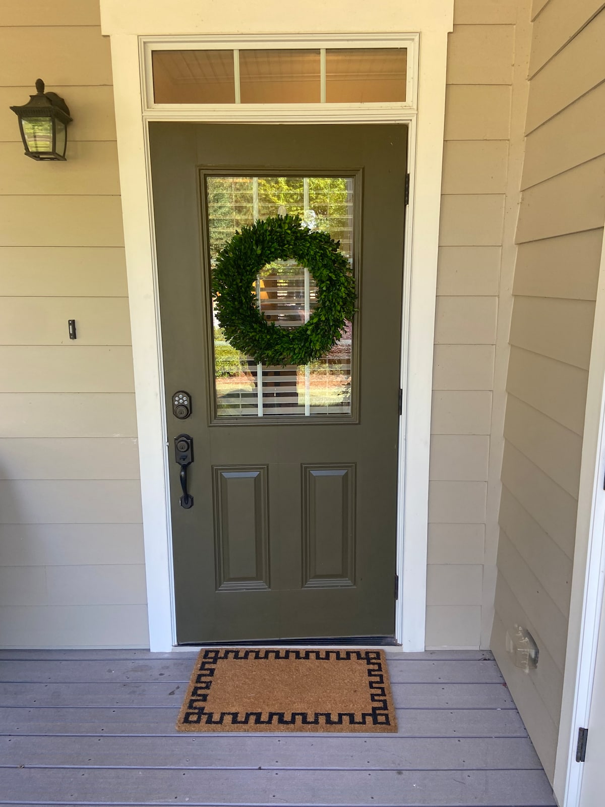 The entrance features a green door adorned with a circular wreath. A welcome mat with a decorative border is placed at the base of the door, while soft natural light filters through the window beside the door.