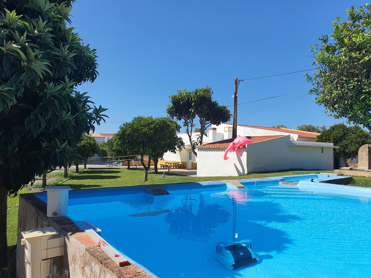 A clear blue pool is surrounded by green grass and trees, featuring a pink float resting on the water. The background includes a single-story house with a yellow roof and a shaded outdoor seating area, enhancing the relaxed setting.