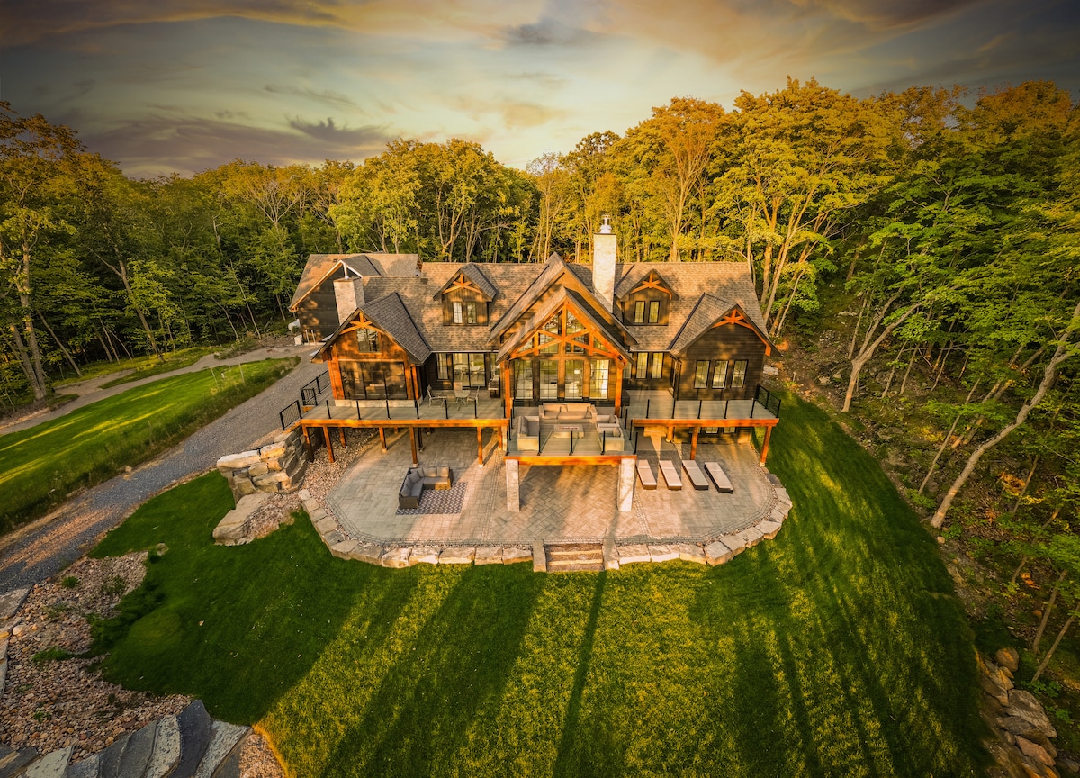 An aerial view of the Sunset Bay Lake House reveals a spacious outdoor deck surrounded by lush greenery. The structure's natural wood exterior harmonizes with the environment, while a stone patio provides ample space for relaxation. Trees line the periphery, enhancing the serene setting.